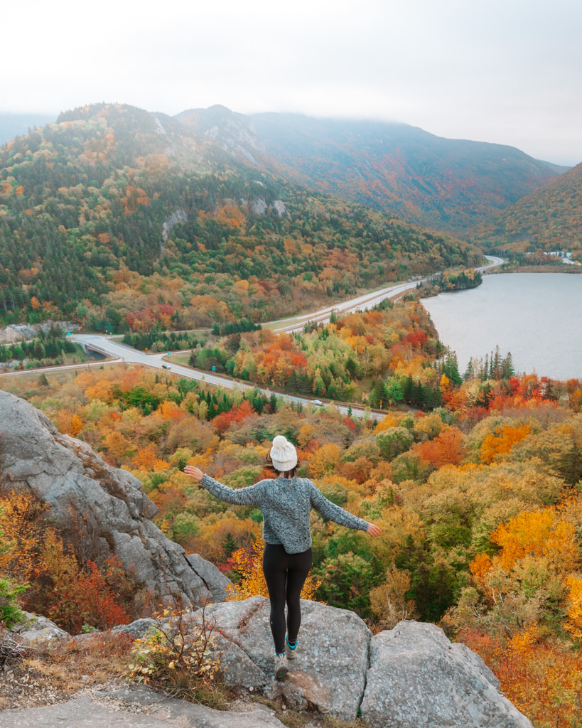 Girl standing on top of Artist Bluff franconia notch state park in fall surrounded by orange and red foliage