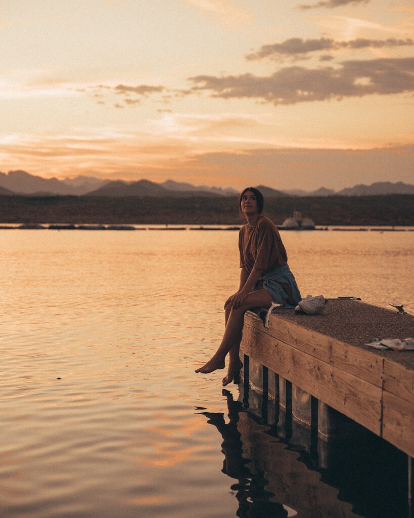 girl sitting on a dock at lake pleasant arizona looking at a super pink sunset with a mountain view