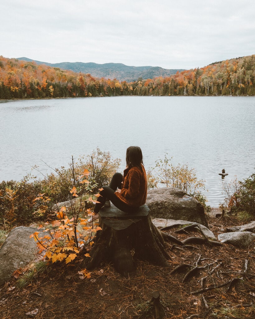 Girl sitting on a tree stump looking at fall colors along the shore of heart lake in the adirondacks ny