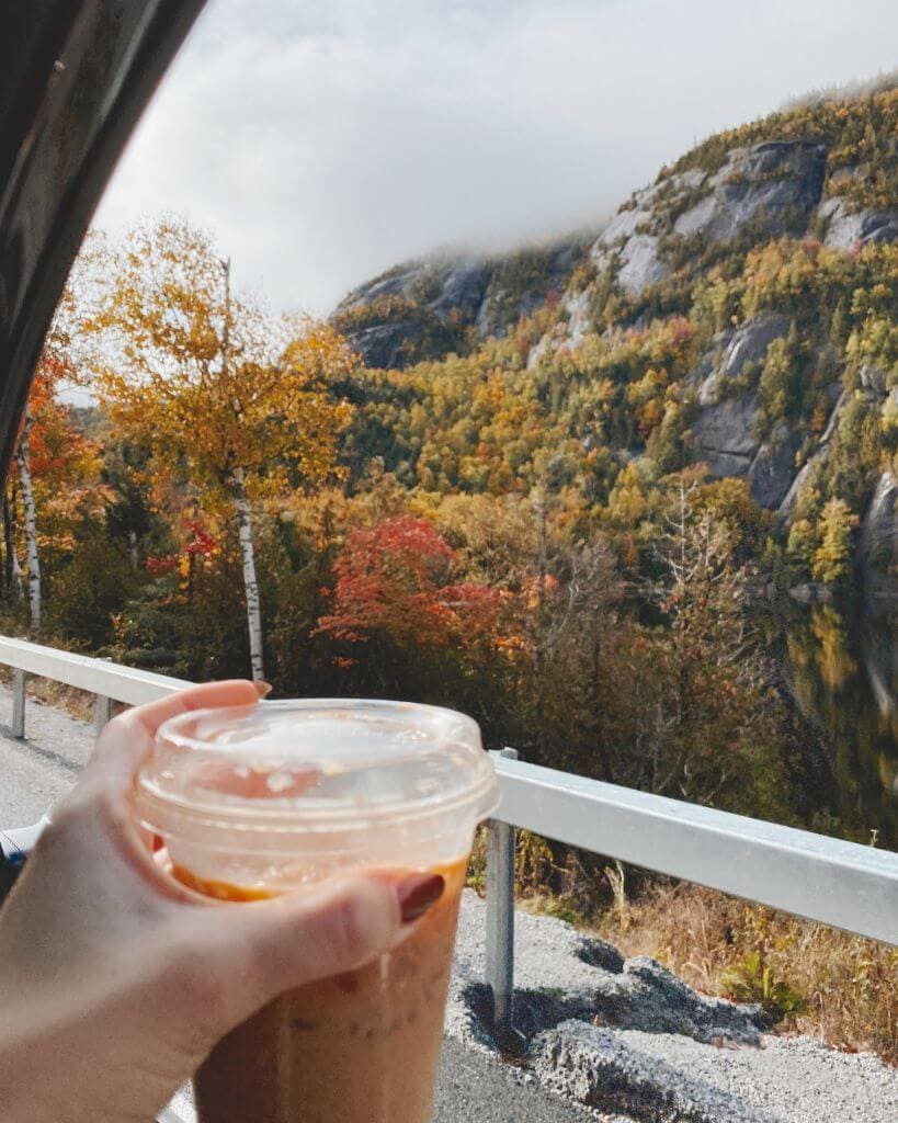 girl holding a coffee cup out a car window while taking a scenic fall drive in the adriondack mountains