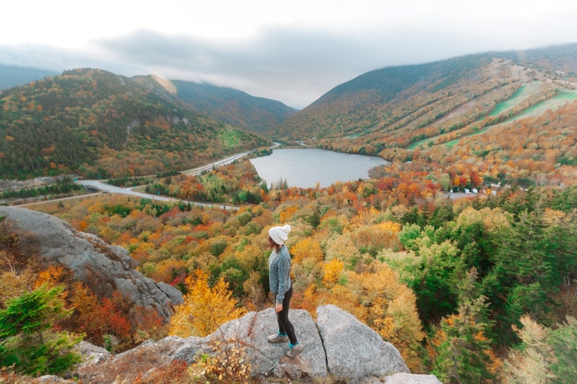 female hiker looking at the orange and gold fall foliage around echo lake over artist bluff nh